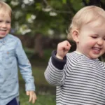 Two young children with blonde hair walking outdoors, surrounded by trees.