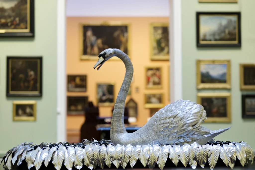 The Silver Swan automaton displayed prominently in a gallery at The Bowes Museum. The swan is crafted from silver with intricate feather detailing and a gracefully curved neck. It rests on a mirrored base surrounded by silver leaves. Behind the swan, the gallery walls are painted pale green and adorned with numerous framed paintings in ornate gold and dark frames. Through an archway in the background, another gallery with peach-colored walls and additional artworks is visible. The wooden floor has a herringbone pattern, and soft lighting highlights the swan’s metallic sheen.