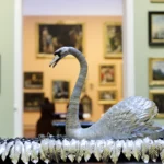 The Silver Swan automaton displayed prominently in a gallery at The Bowes Museum. The swan is crafted from silver with intricate feather detailing and a gracefully curved neck. It rests on a mirrored base surrounded by silver leaves. Behind the swan, the gallery walls are painted pale green and adorned with numerous framed paintings in ornate gold and dark frames. Through an archway in the background, another gallery with peach-colored walls and additional artworks is visible. The wooden floor has a herringbone pattern, and soft lighting highlights the swan’s metallic sheen.