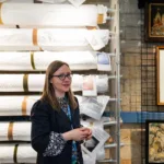 A woman with glasses and long brown hair speaks in front of a storage rack filled with rolled textiles, with framed artwork on the wall behind her in a museum storeroom.