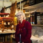 A smiling woman in a red outfit stands in a museum storeroom surrounded by intricately carved antique furniture and ornate wooden cabinets on metal shelving.