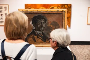 Two women in a museum gallery looking at a Norman Cornish painting on display.