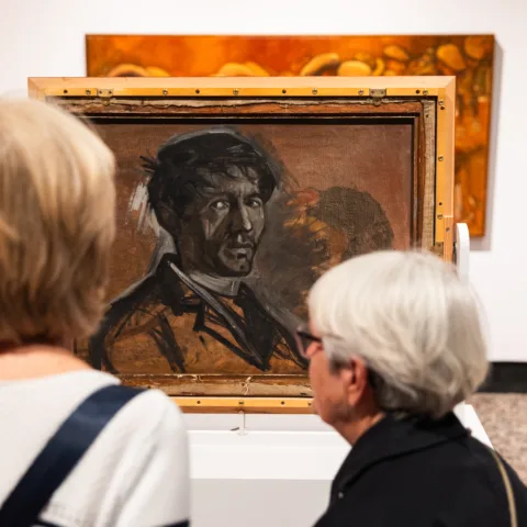 Two women in a museum gallery looking at a Norman Cornish painting on display.