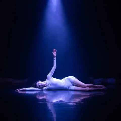 Person dressed in white, laying down on their side on a stage in a pose, under a blue light