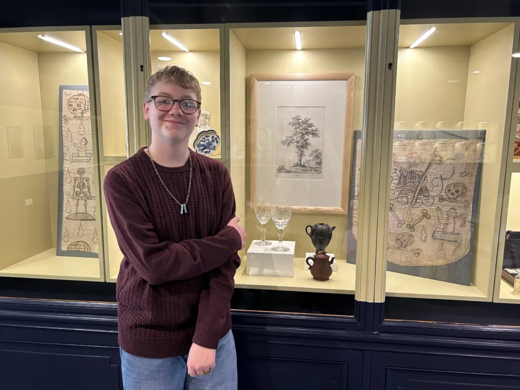 A teenage boy with blonde hair and glasses stands in front of a glass case. The case contains several Museum artifacts.