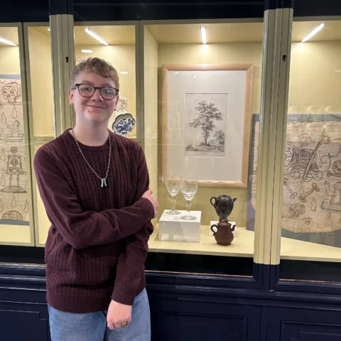 A teenage boy with blonde hair and glasses stands in front of a glass case. The case contains several Museum artifacts.