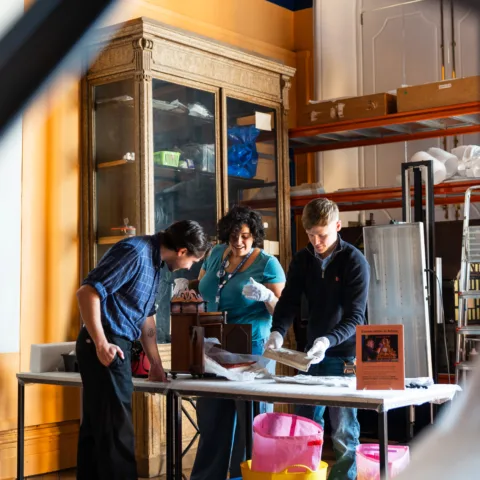 Three people interacting in a museum gallery, looking at a historical object as it's being cleaned for display.