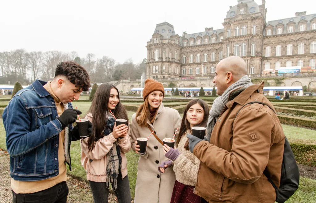 A group of people enjoying hot drinks with striped straws, dressed warmly in winter clothing, smiling and chatting in front of The Bowes Museum, a historic French-style building with gardens in the background.