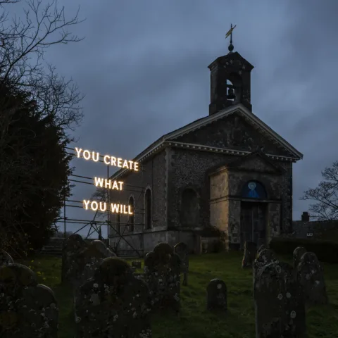 An illuminated text installation reading ‘YOU CREATE WHAT YOU WILL’ in front of a church at night. Gravestones are scattered across the foreground in a grassy churchyard, and bare trees frame the scene.