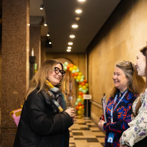 Three woman standing in a corridor in the museum. They are in front of a balloon arch. One is wearing a Bowes Museum lanyard and holds a clipboard.