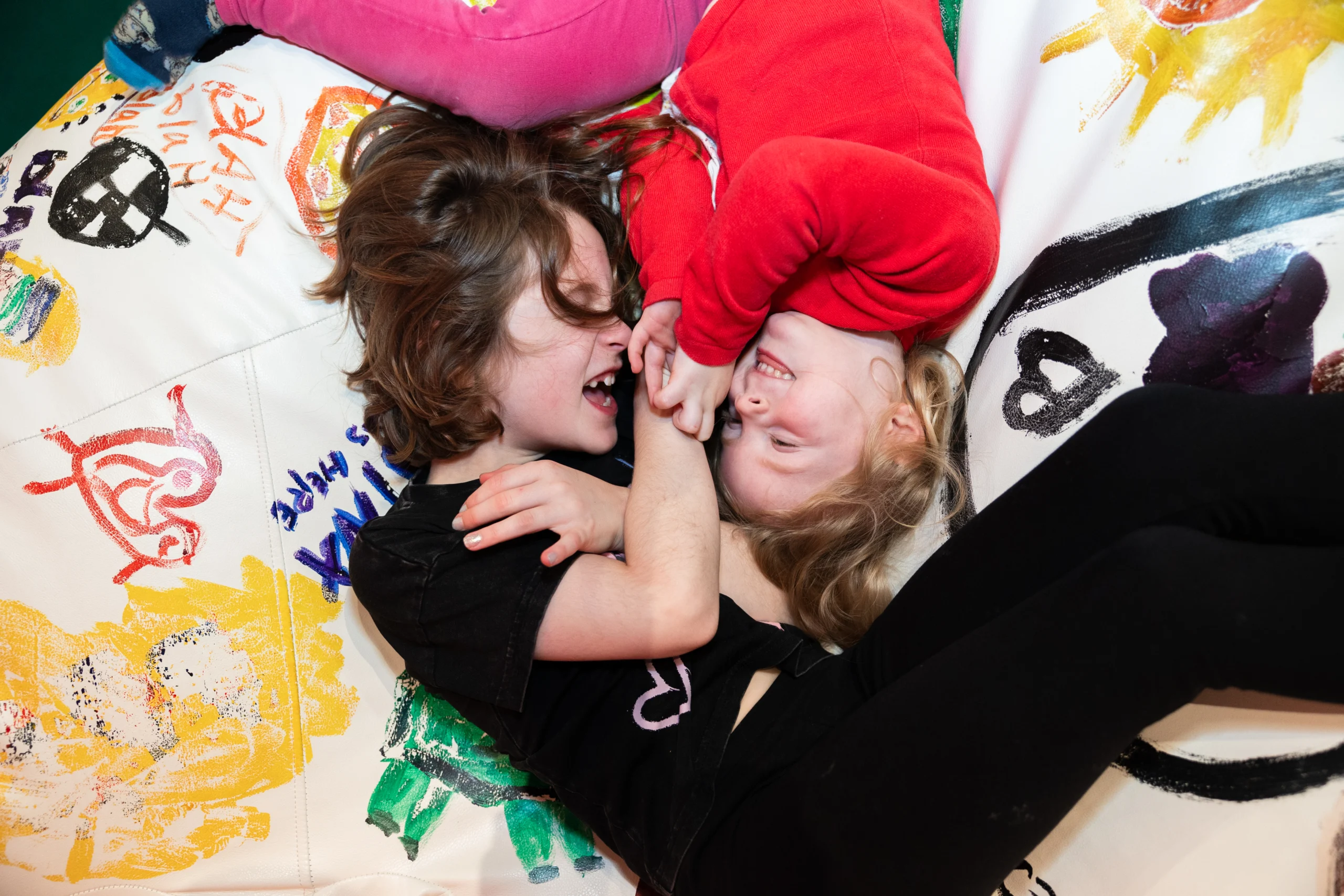Two children lying on a beanbag decorated with colorful artwork, holding hands and resting among other cushions.
