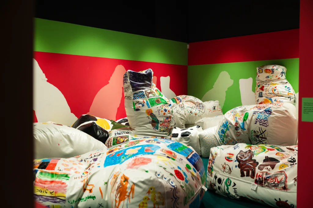A gallery space with green and red walls featuring large beanbag-style cushions covered in colorful, hand-drawn designs and doodles. The cushions are arranged in clusters, creating an interactive and playful environment.