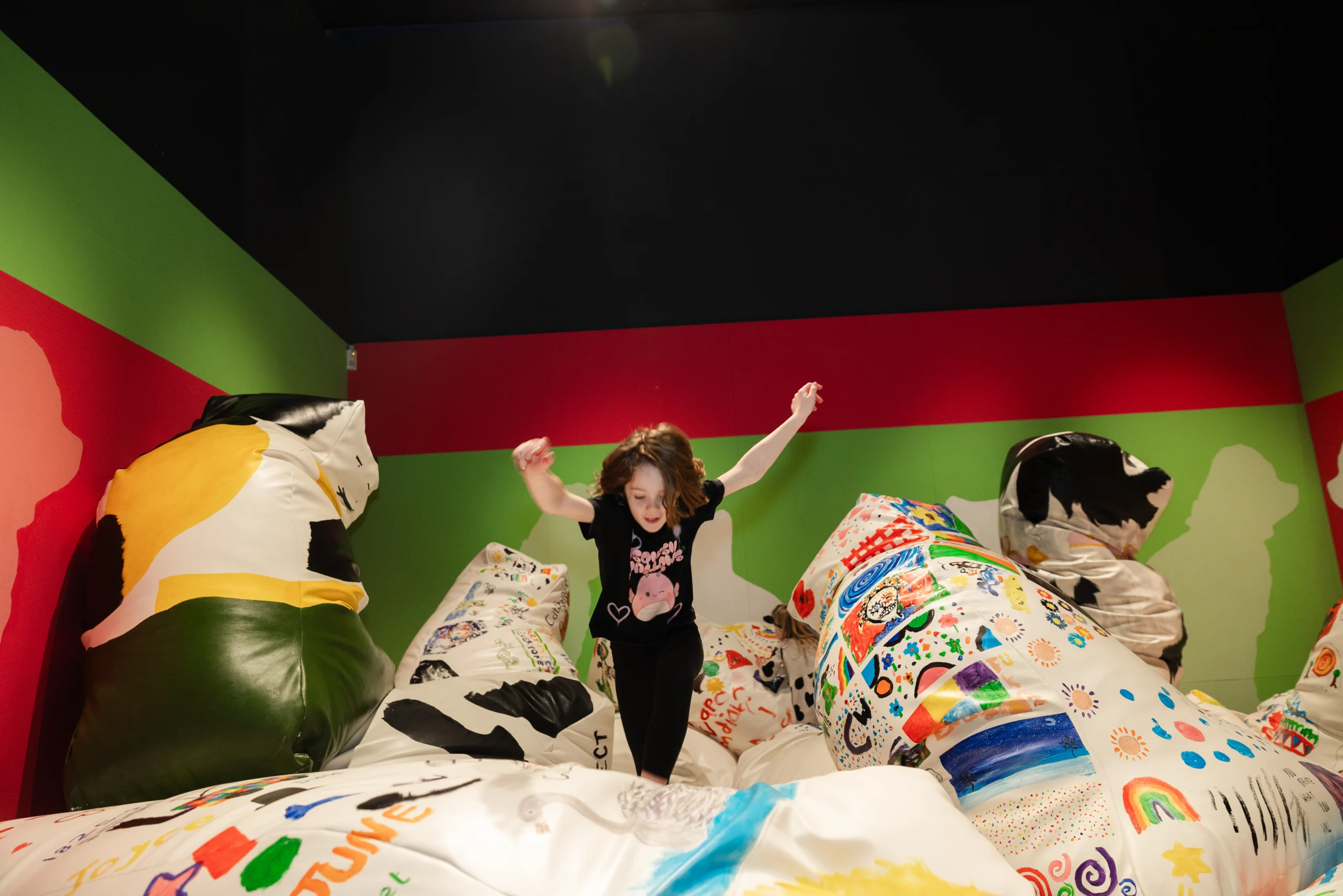 A child standing on a colorful beanbag with arms raised, surrounded by other decorated cushions in a gallery space with green and red walls.