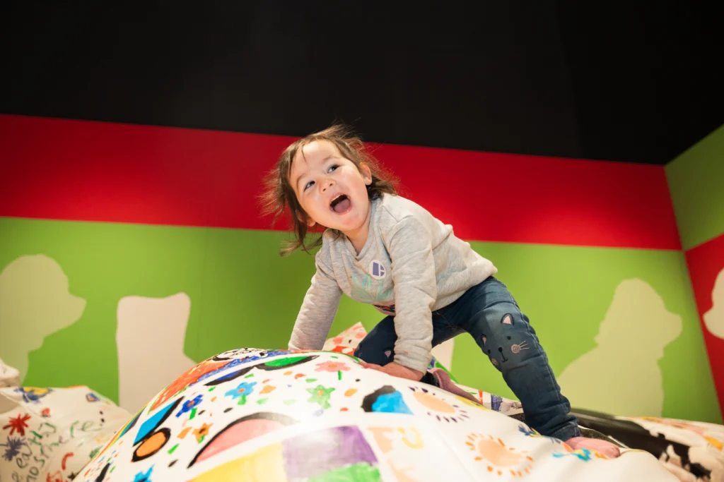 A child climbing on a large beanbag decorated with colorful drawings and patterns in a gallery space with green and red walls.