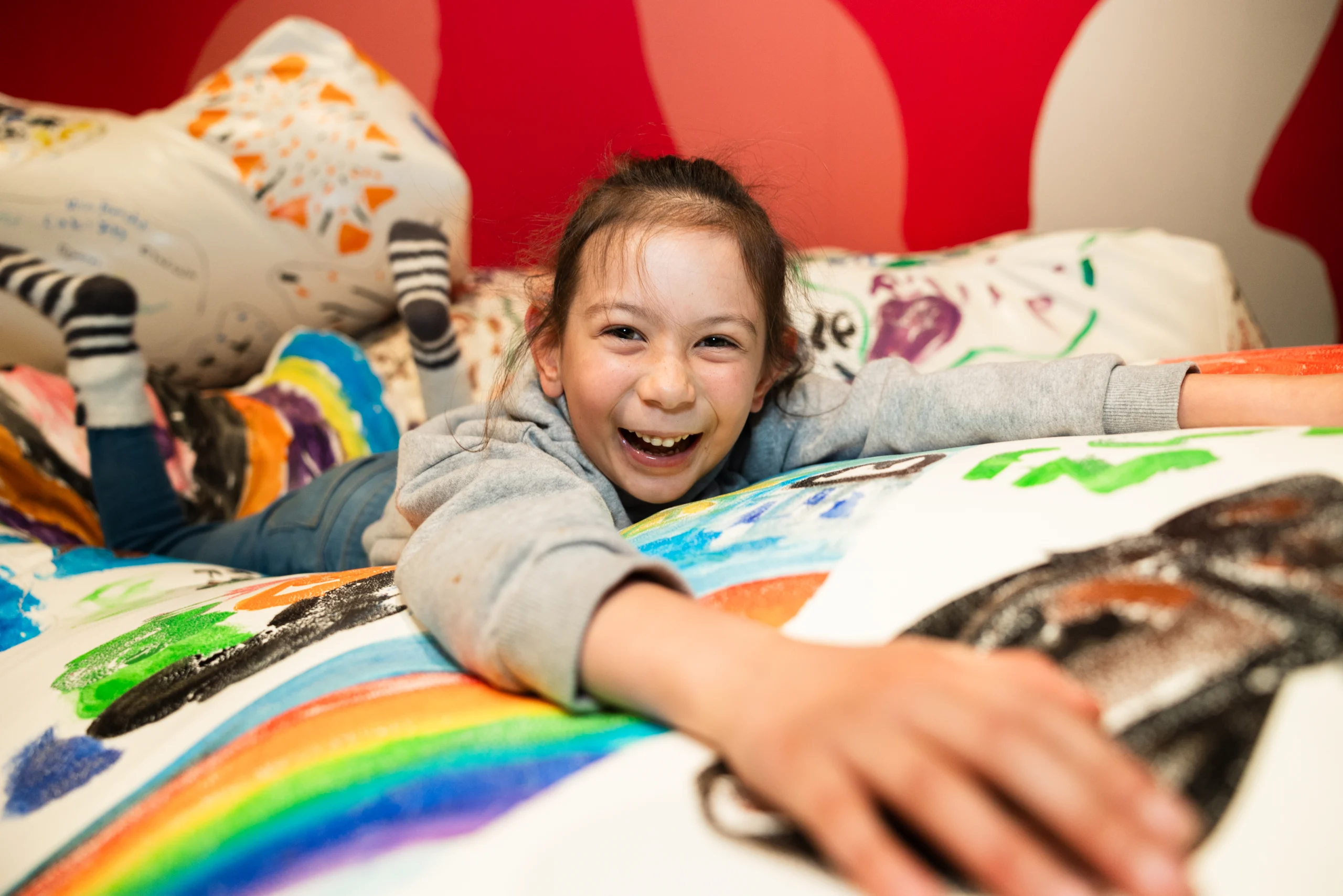 A child lying face down on a beanbag covered in vibrant, hand-drawn artwork, stretching arms forward. The beanbag features rainbows, shapes, and colorful designs.