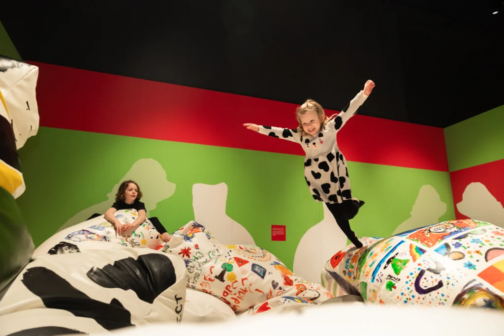 A child in a patterned outfit balancing on a decorated beanbag with arms outstretched, while another child sits on a nearby cushion. The gallery walls are painted green and red.