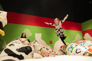 A child in a patterned outfit balancing on a decorated beanbag with arms outstretched, while another child sits on a nearby cushion. The gallery walls are painted green and red.