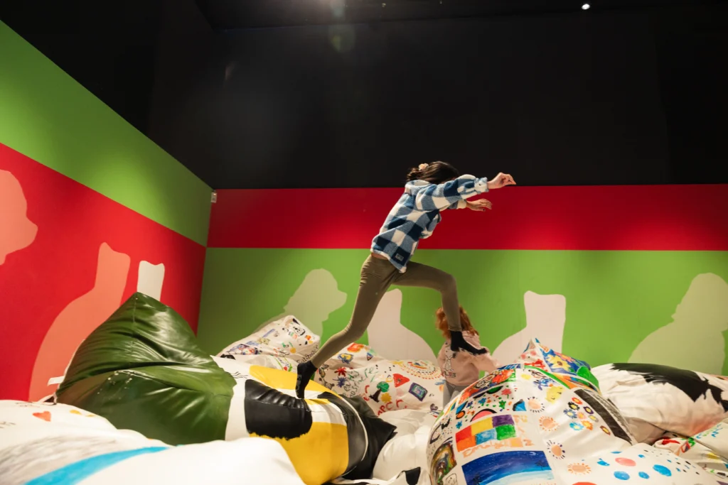 A child mid-jump across large beanbags covered in colorful drawings and patterns, in a gallery with green and red walls.