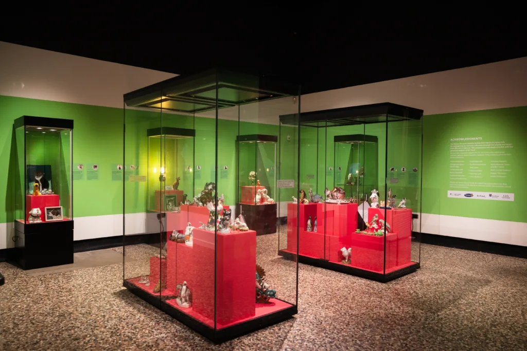 Museum exhibition room with green walls, displaying ceramic figurines in large glass cases with pink platforms inside.