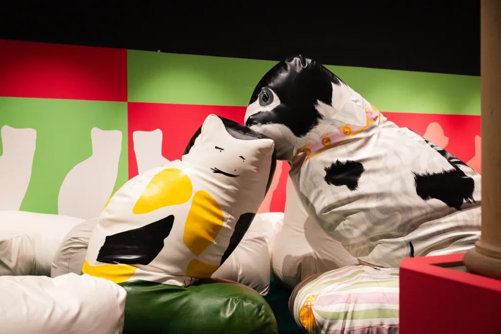 Large soft sculptures resembling a cat and dog with painted features and patterns, displayed against a pink and green wall with cat silhouettes