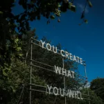 A light installation reading "YOU CREATE WHAT YOU WILL" by Nathan Coley, set against the historic Bowes Museum grounds.