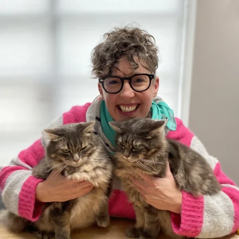 A smiling woman with short curly hair and glasses, wearing a pink and gray sweater and a turquoise scarf, holding two fluffy gray cats on a wooden table.