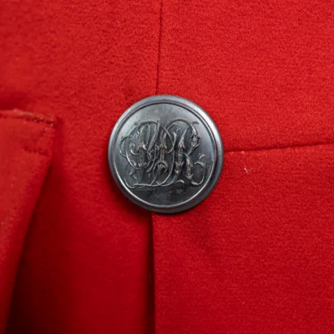 A detail shot of a Guard’s Jacket with a tailored structure, made of red fabric with a metallic button attached. The button has an intricate engraved monogram design.