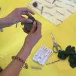 Person shaping clay to make a shaped filled with seeds at a craft table with tools, tins and a seed packet.