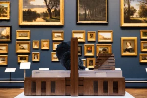 Nathan Coley’s sculptural model of the Tate Modern with added plumes of dark smoke, giving the illusion of the building on fire. The piece is displayed on a white table in a gallery room lined with traditional landscape and portrait paintings.