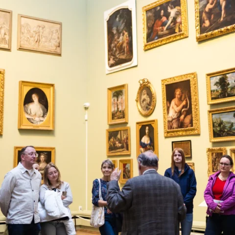 A group of people listening to a guide in an art gallery, surrounded by a collection of framed paintings and sketches on a green wall.