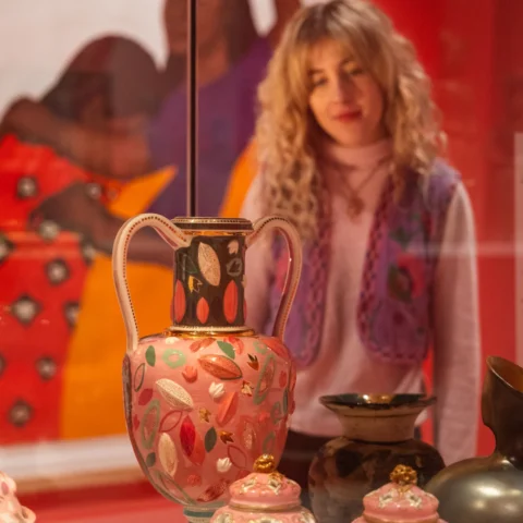 A woman with curly blonde hair wearing a pink sweater and a purple embroidered vest observes a display of ornate ceramic vases in a museum, with a colourful painting of figures in the background.