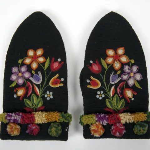 A pair of black wool mittens with vibrant floral embroidery in red, orange, purple, and green, featuring a textured, multicoloured cuff with small pom-poms.