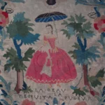 An embroidered textile chair back, featuring a central figure of a woman in a pink dress holding a blue parasol, surrounded by floral motifs, trees, and the French phrase 'La beauté nuit à la pudeur' stitched below.