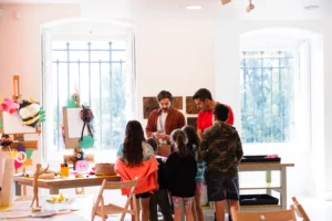 A group of children and an adult gather around a male adult instructor in a bright, art-filled classroom, watching attentively as he demonstrates a craft project.