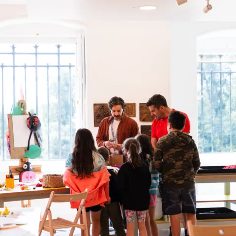 A group of children and an adult gather around a male adult instructor in a bright, art-filled classroom, watching attentively as he demonstrates a craft project.