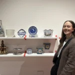 A young woman with long brown hair, wearing a plaid blazer, smiles while standing beside a display of decorative historic and contemporary ceramics on white shelves, including a fish-shaped jug, blue and white plates, and ornate porcelain pieces.