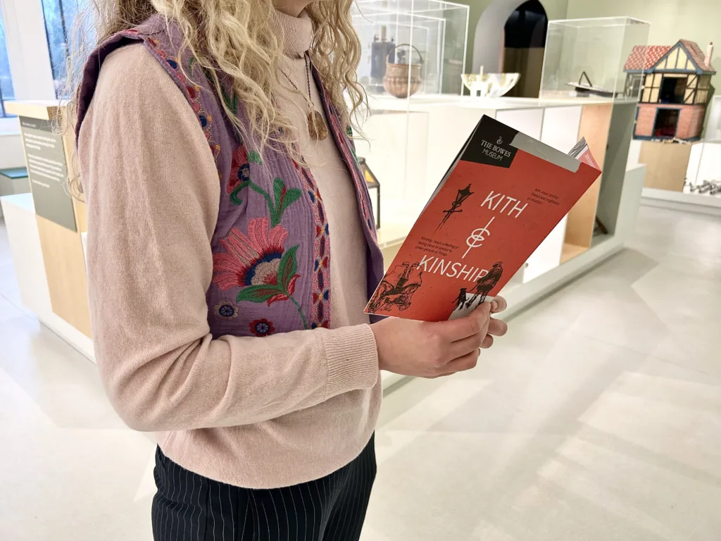 A person in a pink turtleneck and embroidered vest holds a red booklet titled 'Kith & Kinship' inside a museum with glass display cases and historical artifacts.