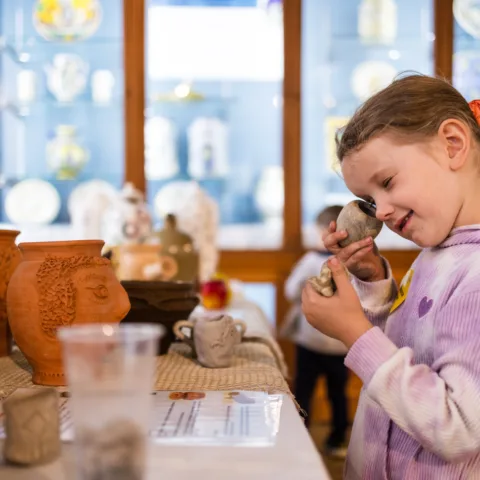 A young girl in a purple tie-dye hoodie looking at a small clay object closely in a object handling workshop, surrounded by handmade pottery and a glass display case filled with decorative ceramics in a gallery.