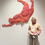 Daisy stood in front of her artwork. She is wearing a pink jumper and green trousers.