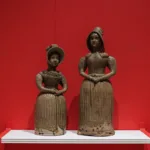 Two ceramic spirit flasks shaped like women in 19th-century attire stand on a white shelf against a vivid red wall, showcasing intricate detailing in their dresses and bonnets.