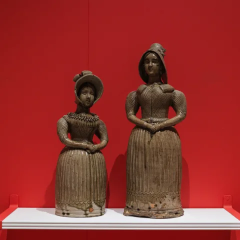 Two ceramic spirit flasks shaped like women in 19th-century attire stand on a white shelf against a vivid red wall, showcasing intricate detailing in their dresses and bonnets.