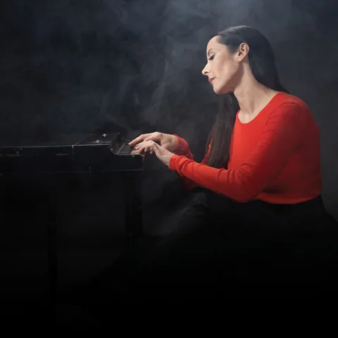 Nerina Pallot sitting at a piano, wearing a red shirt. She is surrounded by mist.