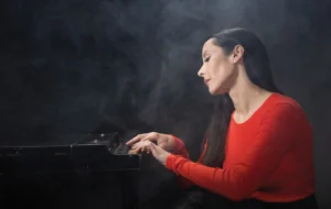 Nerina Pallot sitting at a piano, wearing a red shirt. She is surrounded by mist.
