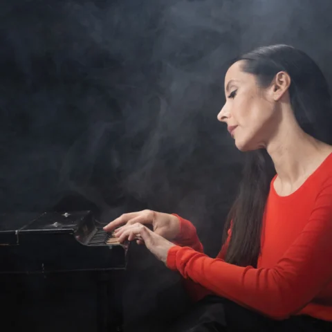 Nerina Pallot sitting at a piano, wearing a red shirt. She is surrounded by mist.