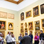 A group of ten people listening attentively to a curator in a museum gallery filled with framed historical paintings.