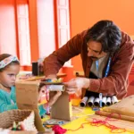A girl making art at a table with help from a young man.
