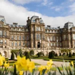 A grand historic building with ornate architecture, large windows, and a landscaped garden with blooming yellow daffodils in the foreground.