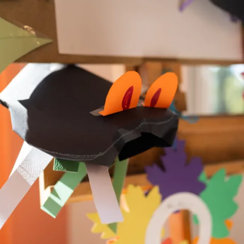 A colourful paper craft resembling a whimsical creature with orange eyes, black body, and green and white legs is displayed among other vibrant paper decorations.