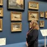 A woman looking at paintings in a gallery. The gallery has dark blue walls. She is focused on a frame that is black, with a painting of a seascape.