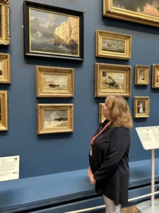 A woman looking at paintings in a gallery. The gallery has dark blue walls. She is focused on a frame that is black, with a painting of a seascape.